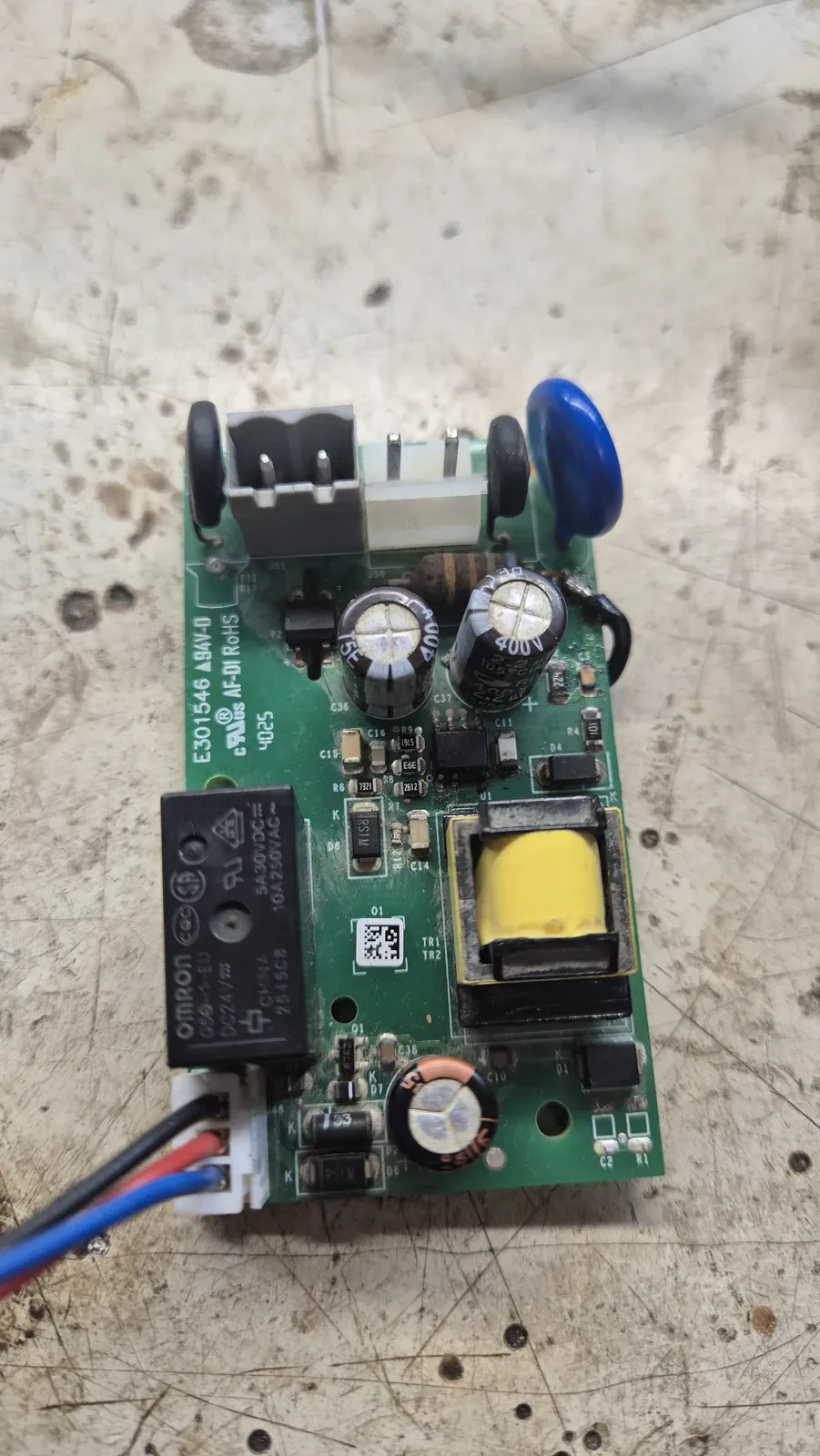 Una scheda elettronica verde con morsetti, relè, condensatori e un piccolo trasformatore, appoggiata sul banco